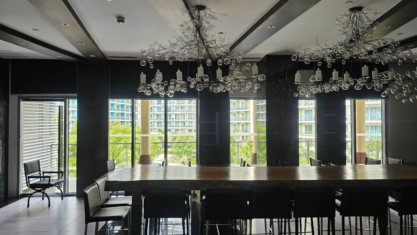 A modern conference room with a long table, black chairs, large windows overlooking buildings and greenery, and two ornate chandeliers hanging from the ceiling.