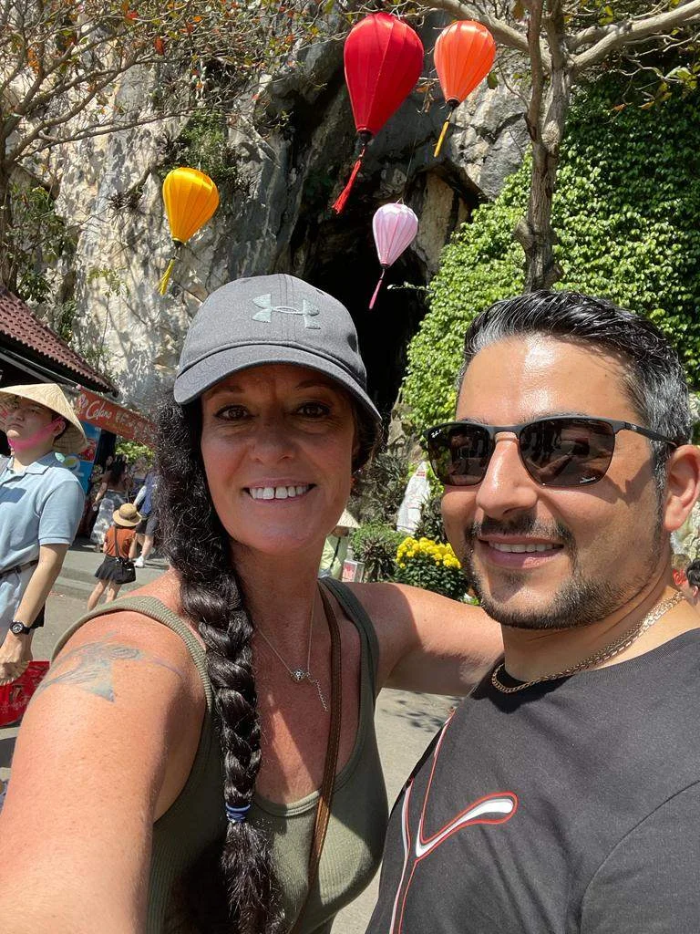 Two people smiling outdoors. The woman on the left wears a cap and tank top, the man on the right wears sunglasses. Balloons float in the background.