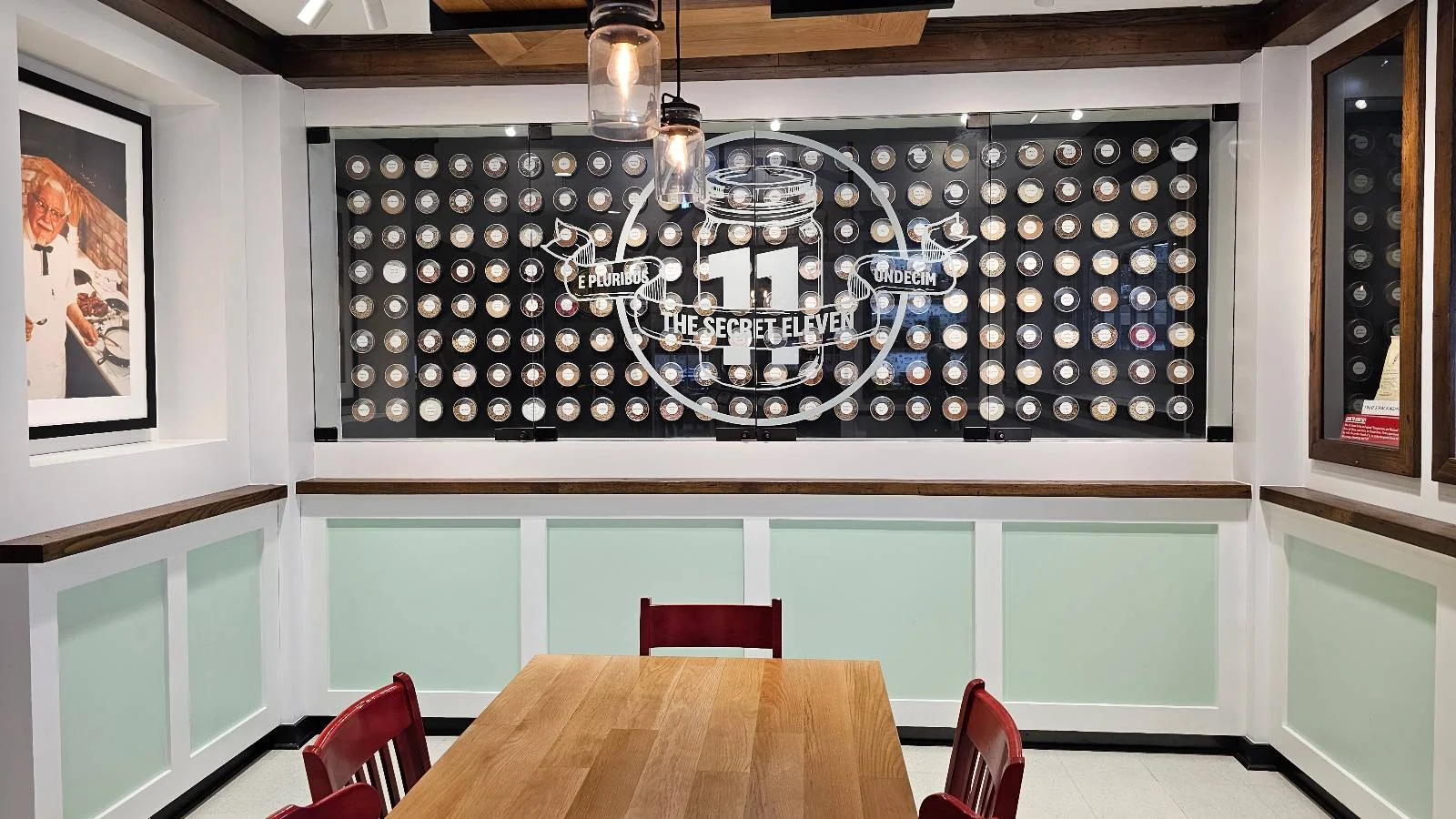 A small dining area with a wooden table and red chairs sits in front of a wall decorated with a large "11 spices" logo and a display of many white spice jars arranged in a grid.