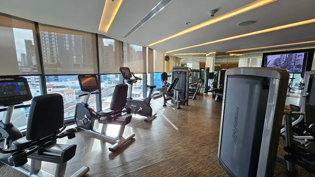 Cardio and strength-training equipment beside floor-to-ceiling windows overlooking Bangkok.