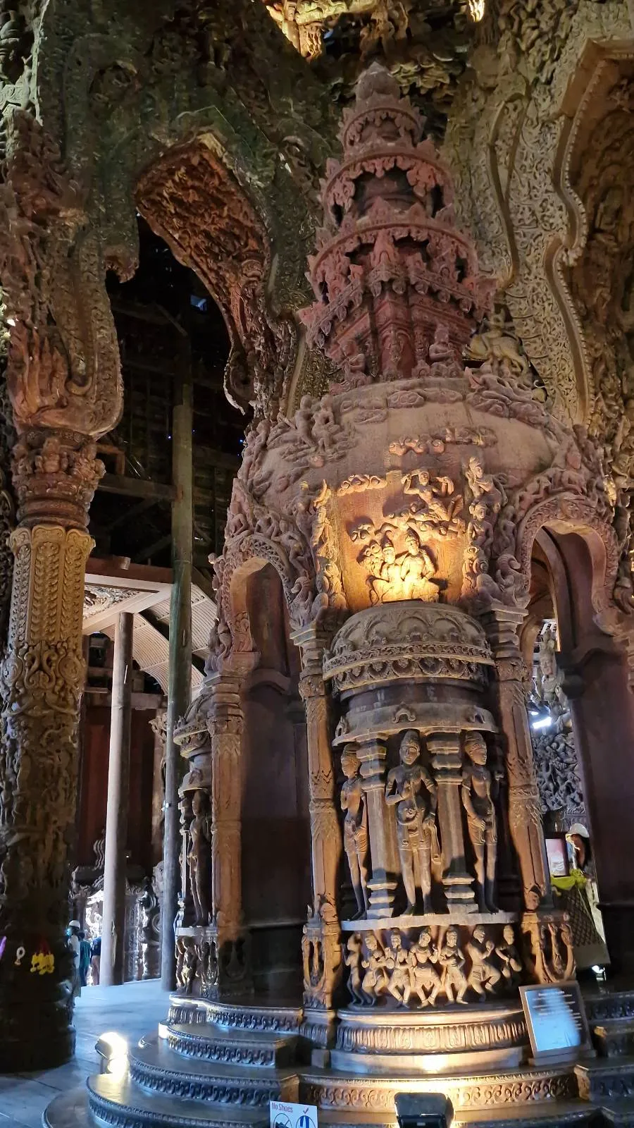 Ornately carved wooden pillars and a central sculpture in a temple interior, featuring intricate designs and figures under a decorative canopy. The scene is dimly lit, highlighting the craftsmanship and intricate details of the carvings.