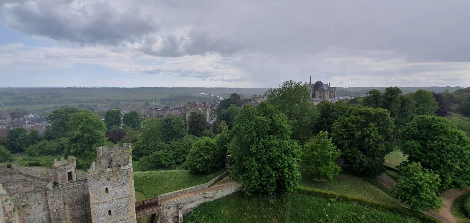 A vast landscape view from a castle, featuring lush green trees and pathways. The sky is cloudy, and a distant town is visible under a broad horizon. Part of the castle structure is on the left.