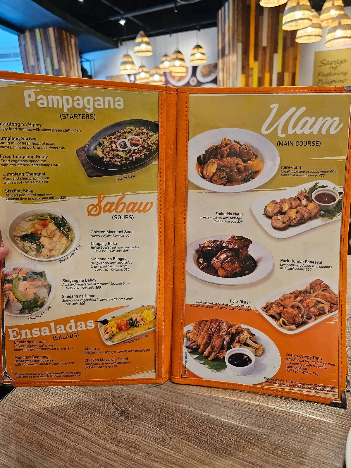 A restaurant menu featuring Filipino dishes. The left page includes Pampagana appetizers, Sabaw soups, and Ensaladas salads. The right page showcases various Ulam main courses with images of each dish. The background shows a decorated dining area.