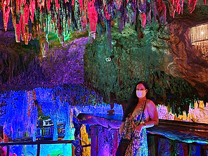 A woman wearing a mask stands beside a wooden railing inside a colorful cave in Mexico. The cave walls and ceiling are illuminated with vibrant lights in pink, green, and blue, highlighting the stalactites and stalagmites, reminiscent of ancient ruins bathed in ethereal hues.