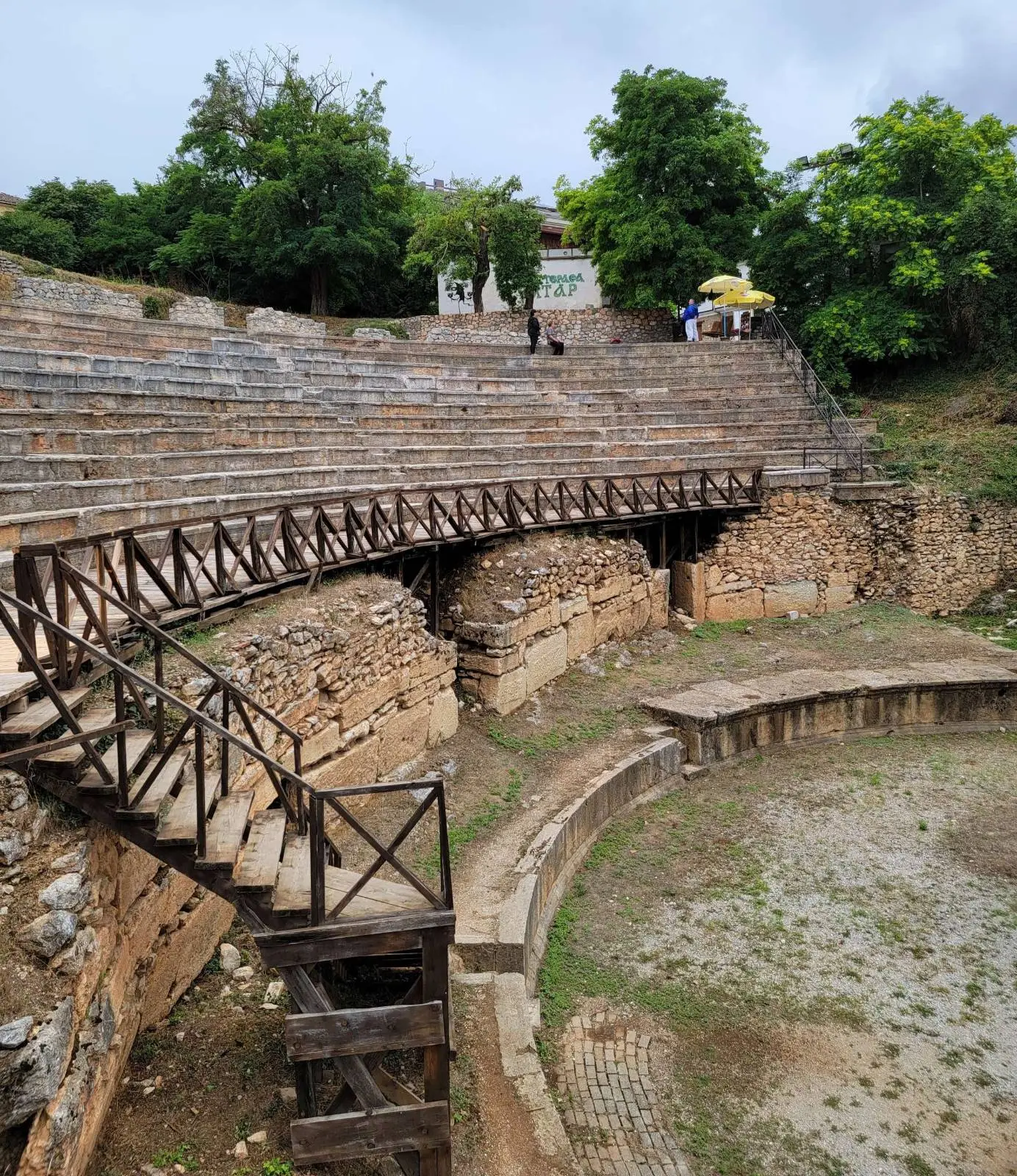 Ancient stone amphitheater with wooden stairways, curved seating, and a partially overgrown stage area, surrounded by trees and a cloudy sky.