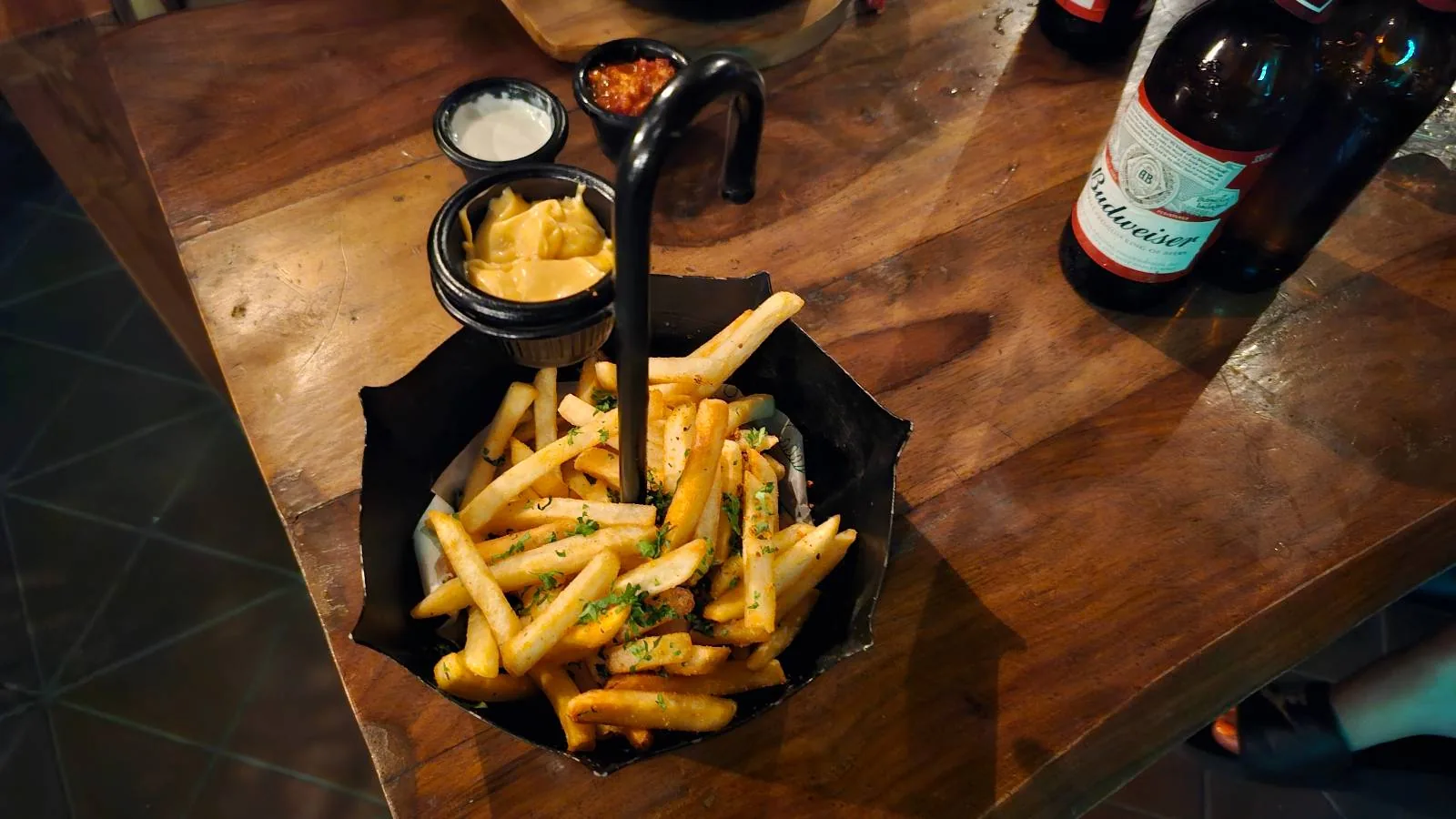 A creative serving of fries placed in an open black umbrella. The fries are garnished with herbs and accompanied by small bowls of dipping sauce. Nearby, there's a bottle of beverage on the wooden table.