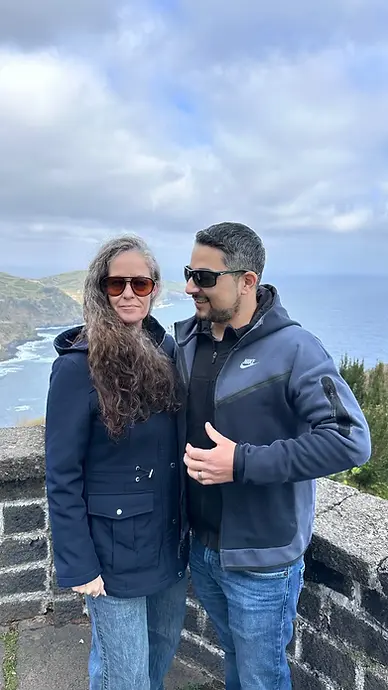 A couple stands together by a stone wall overlooking the ocean and rocky coastline. Both wear jackets and sunglasses, with cloudy skies above and green hills in the background.