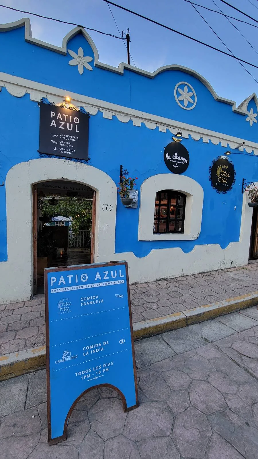 The image shows a vibrant blue building with a sign that reads "Patio Azul." The facade features decorative circular signs and a menu board on the sidewalk. The architecture is traditional with arched doorways and windows.