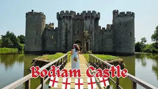 Woman in a blue dress walks on a bridge towards Bodiam Castle over a moat, with "Bodiam Castle" text at the bottom with red and white flags.