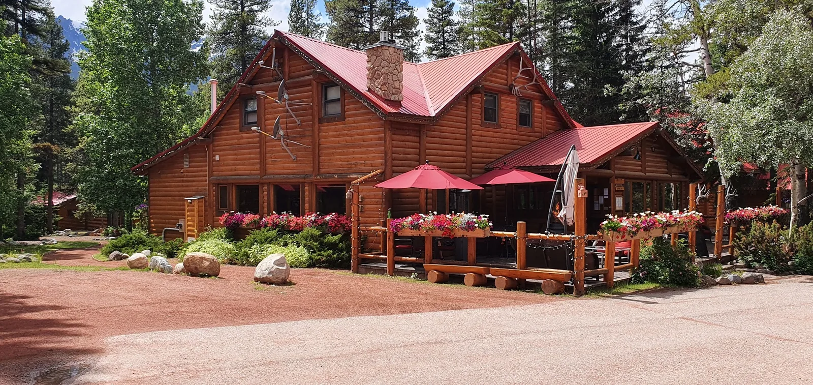 A rustic wooden cabin with a red roof, surrounded by trees. The porch has red umbrellas and potted flowers.