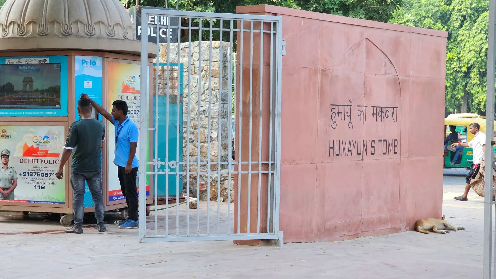 People stand outside a stone and brick building with open doorways. A dog lies on the ground in the foreground. Trees are in the background and a colorful sign is on the right. It's a sunny day.
