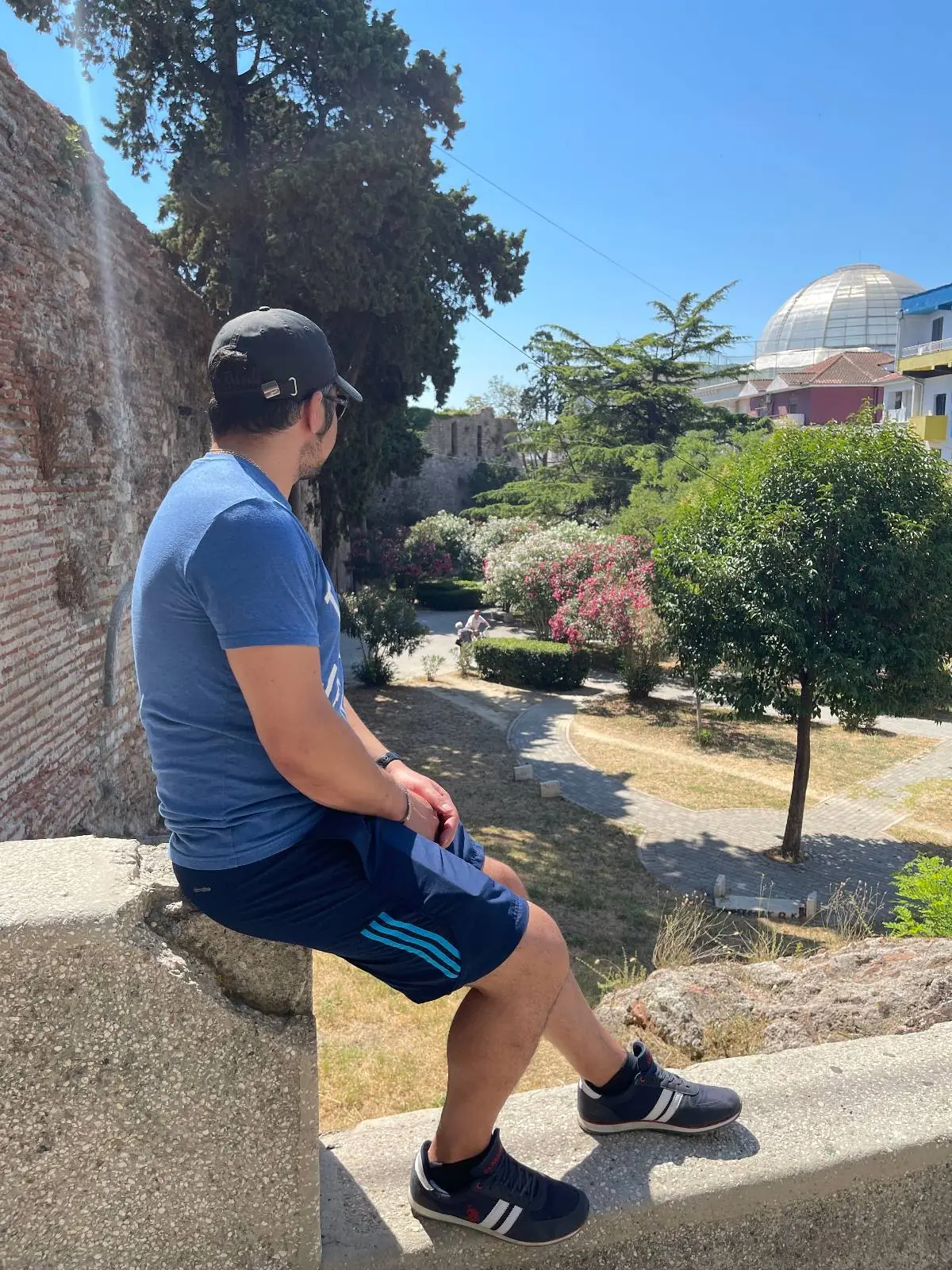 A woman wearing sunglasses, a gray cap, and a black and yellow swimsuit poses for a selfie outdoors. Behind her are potted plants, a building, and sunny weather.