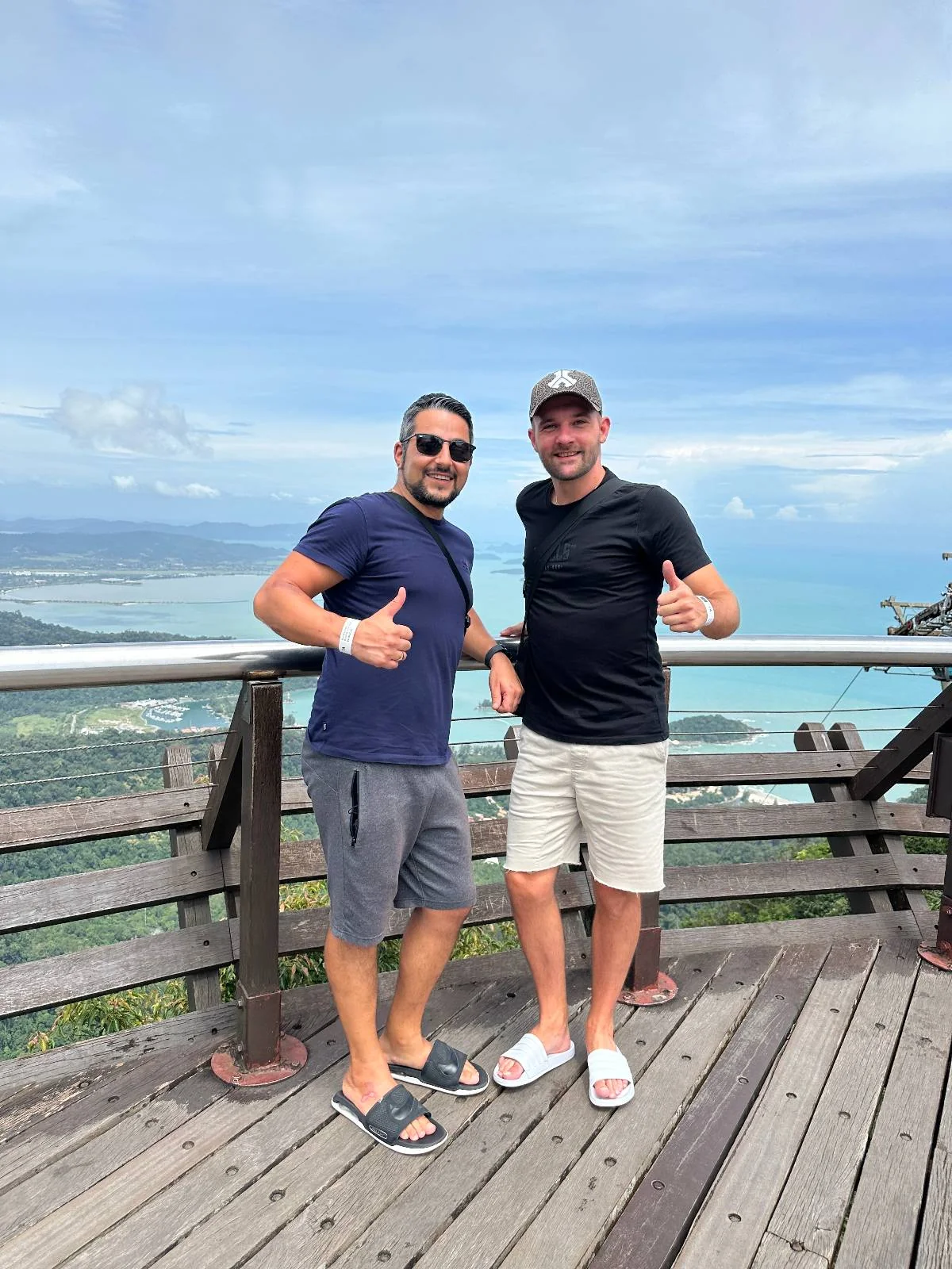 Two men are standing on a wooden deck with a scenic ocean view behind them. Both are smiling, wearing casual summer clothes and sunglasses. One man wears a cap, and they both have their thumbs up, enjoying the day.