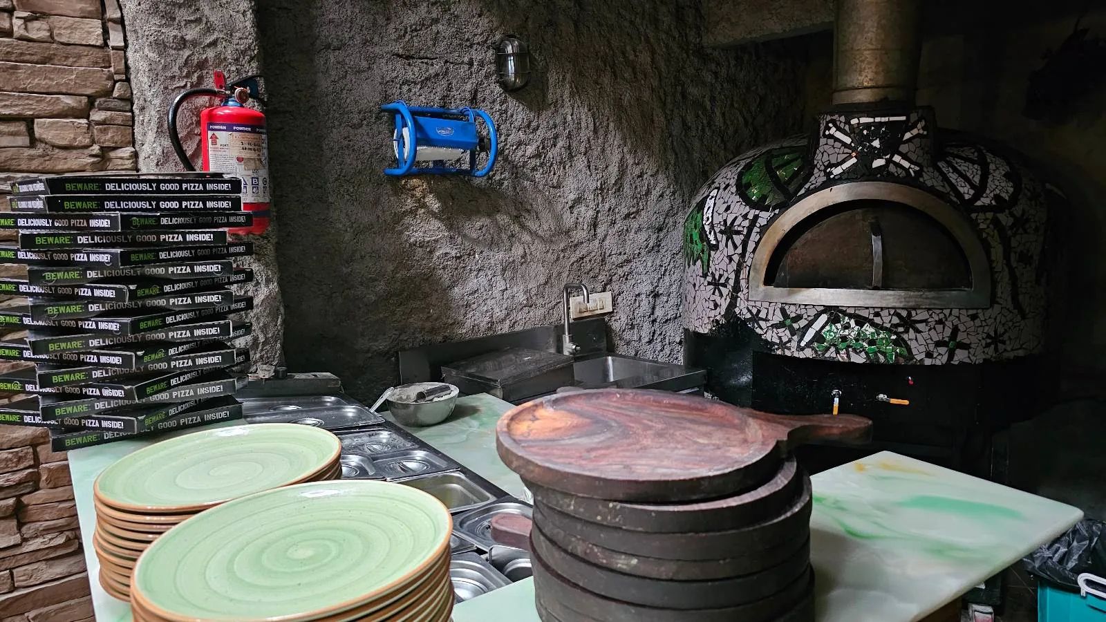 A rustic kitchen with a stone pizza oven adorned with artistic designs. Stacks of plates and trays are on the counter. Pizza boxes are neatly piled on one side, and a fire extinguisher is mounted on the wall.