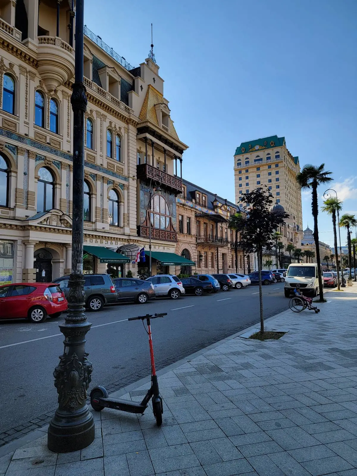 A street scene with historic buildings, parked cars, and a row of trees. A black and red electric scooter is parked by a lamppost on the sidewalk. A tall modern building is visible in the background under a clear blue sky.