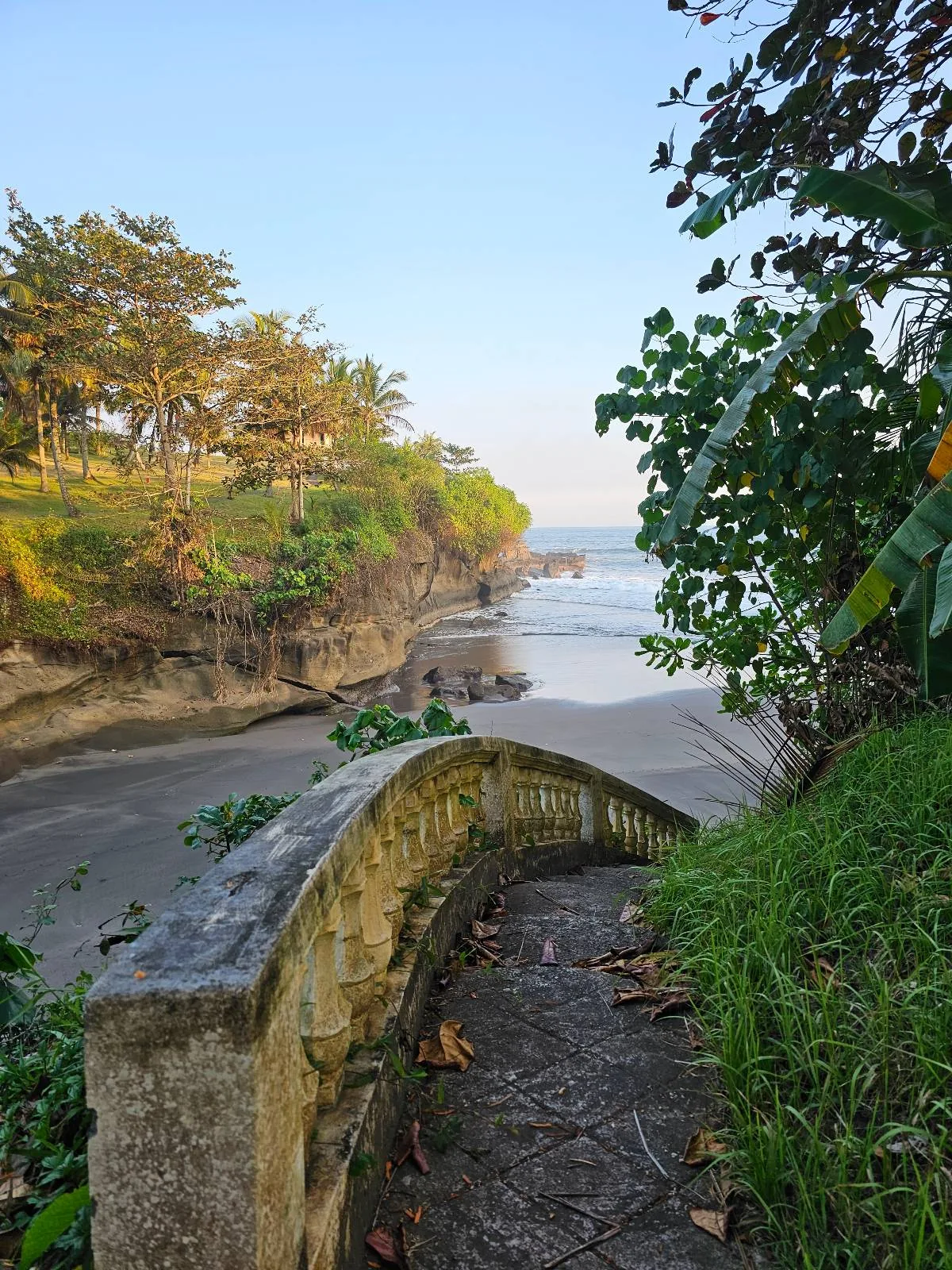 A stone pathway curves along the edge of a lush, green cliff overlooking a secluded sandy beach. The ocean is calm with gentle waves, and trees line the top of the cliff under a clear blue sky.