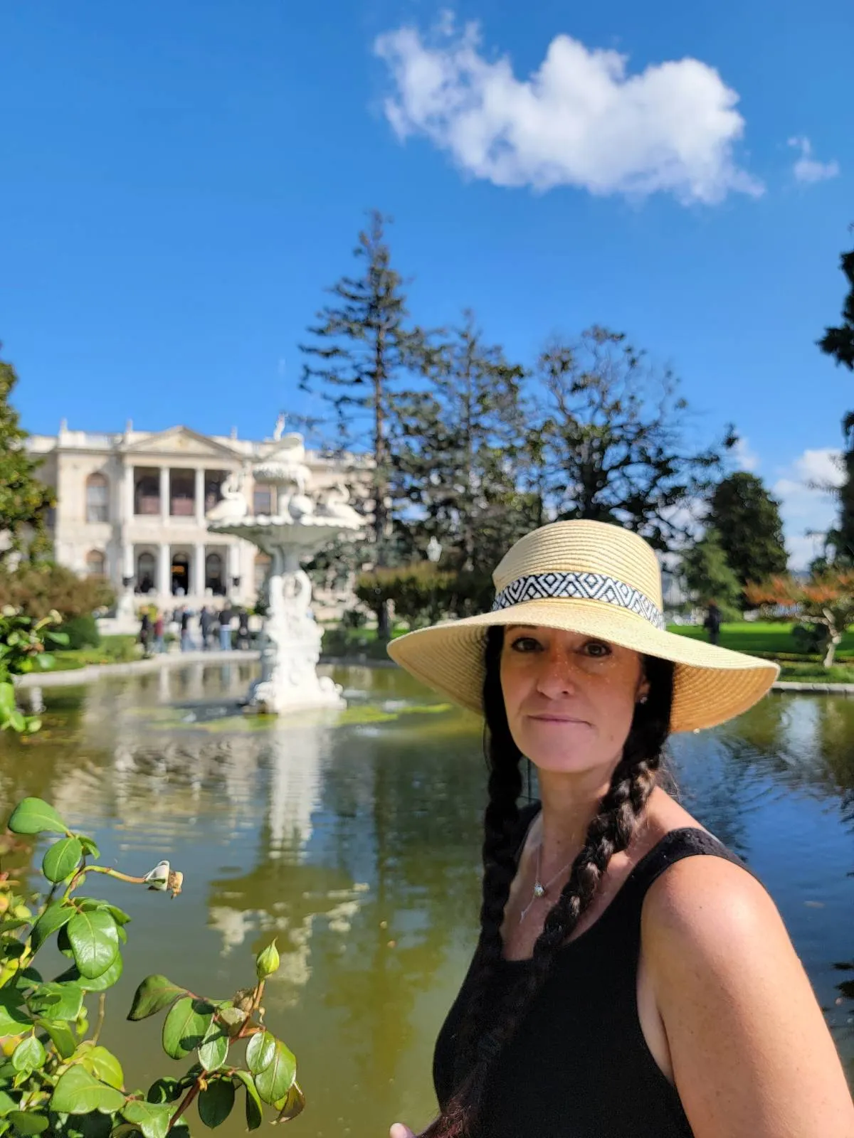 A woman with a sun hat stands in front of a pond with a statue, and a mansion is visible in the background under a clear blue sky.