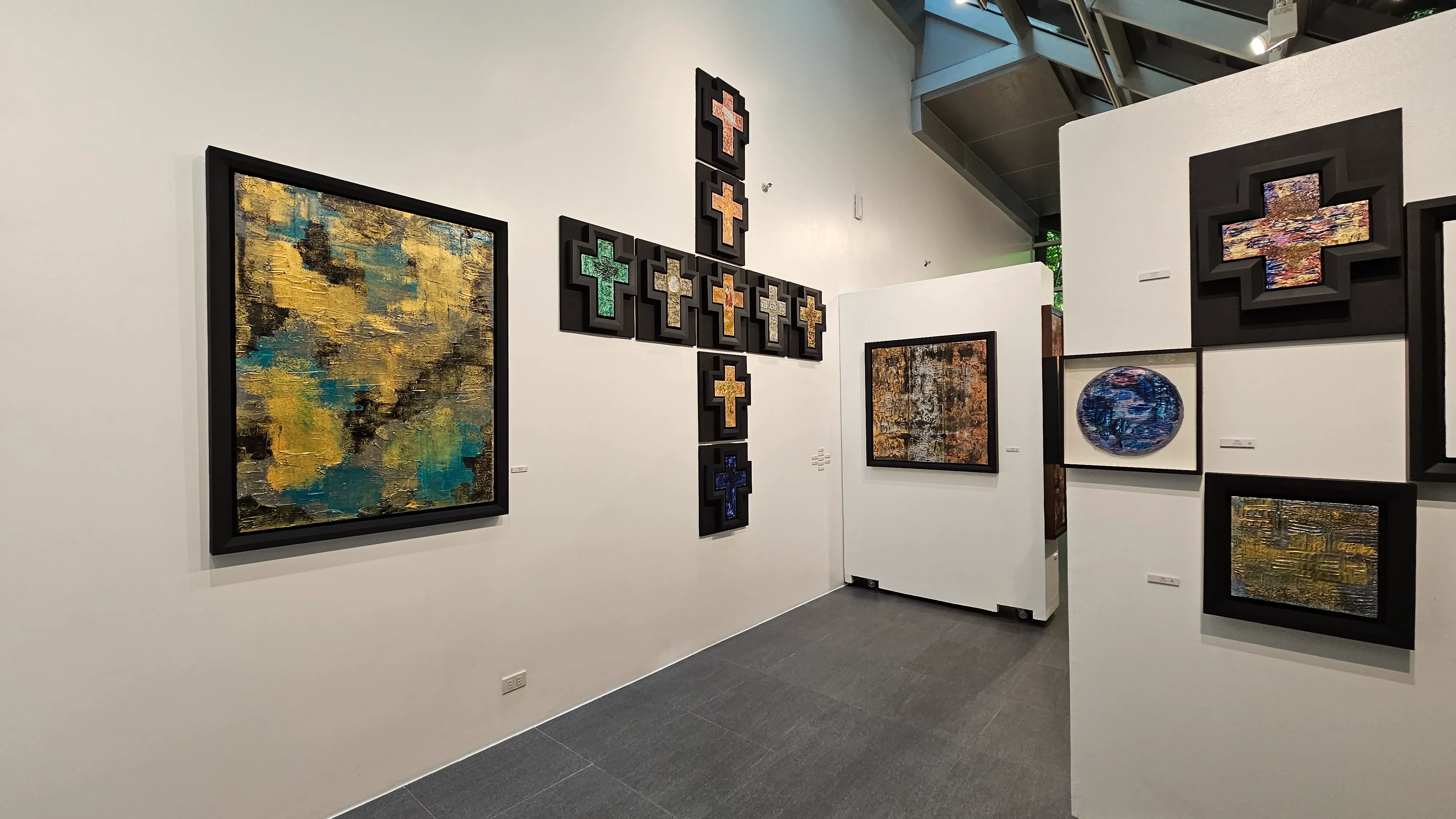 Art gallery interior with various framed artworks on white walls, including abstract paintings, cross-shaped pieces, and a circular blue artwork, all displayed in a well-lit, modern exhibition space.