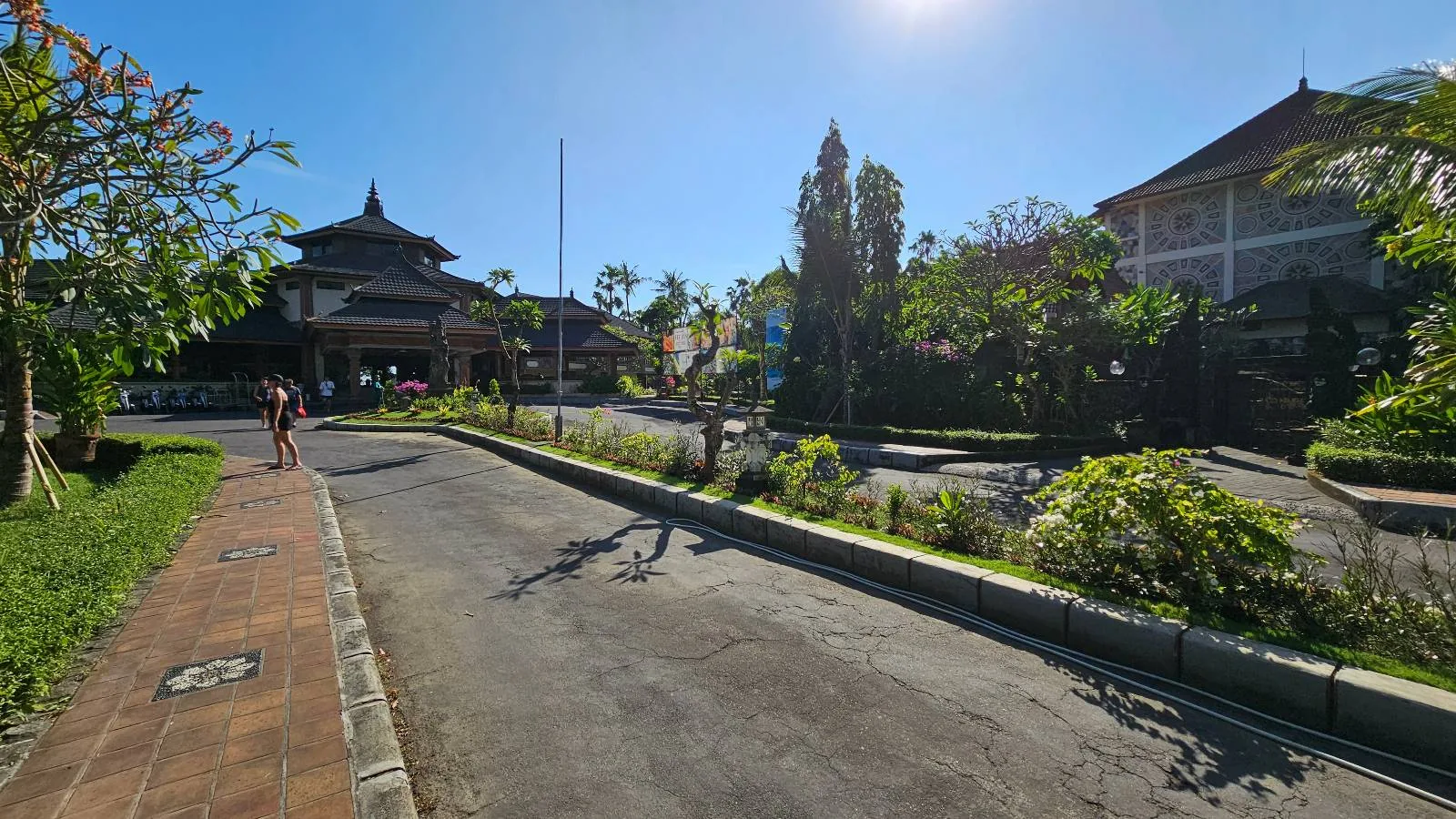 A paved road with a cyclist in the distance passes by lush greenery and traditional, pagoda-style buildings under a clear blue sky. Sunlight casts long shadows on the ground, highlighting the serene and sunny atmosphere.
