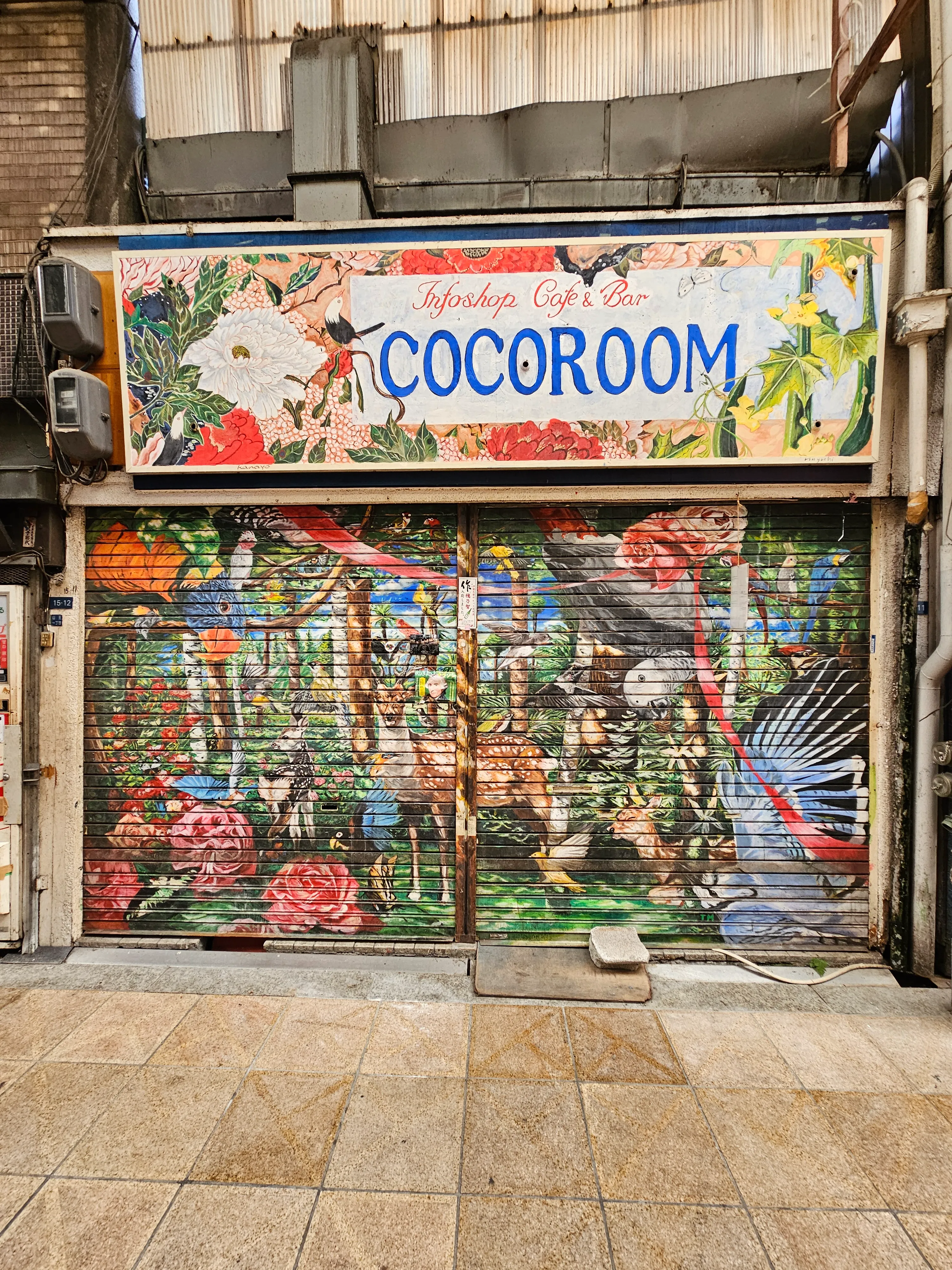 A shopfront with a sign reading "COCOROOM" above a colorful mural featuring flowers, birds, and lush plants. The artwork covers the entire closed shutter, giving the area a lively, vibrant appearance.