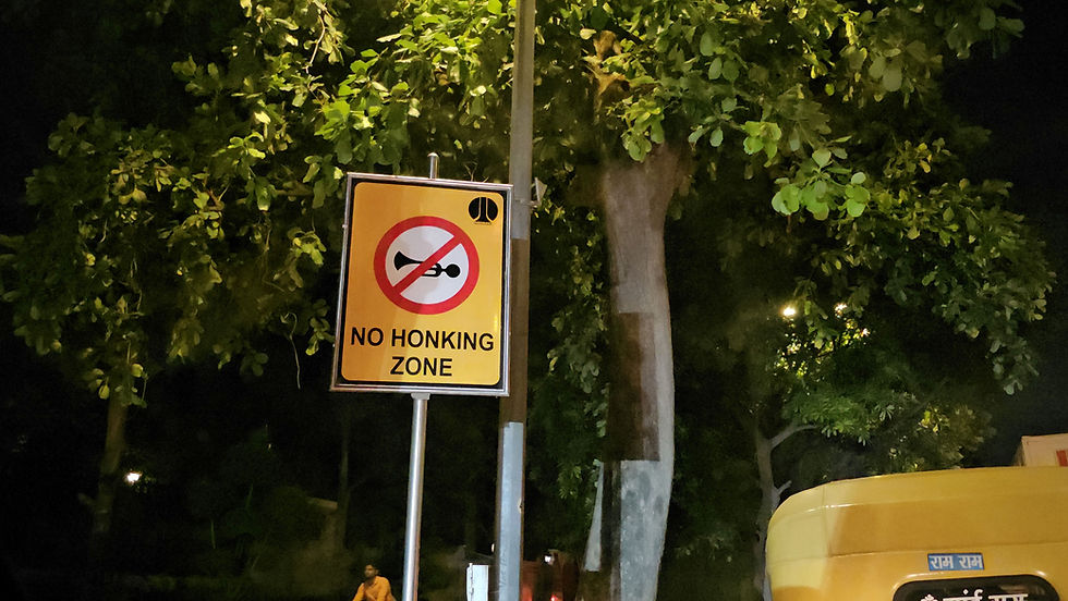 A "No Honking Zone" sign on a pole at night, surrounded by trees. A yellow vehicle with text in a script visible below. Calm ambiance.