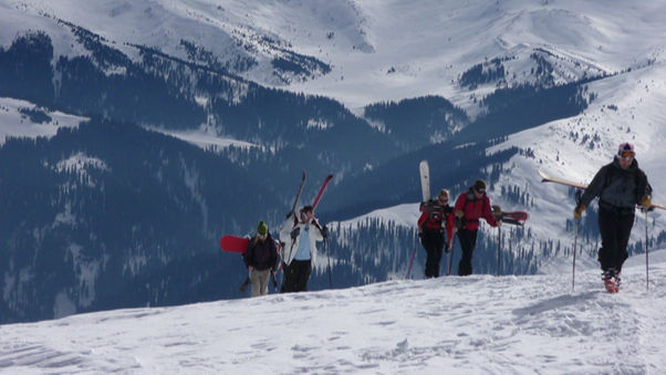Skiers carrying gear hike up a snowy mountain slope. The backdrop features snow-covered peaks and dark tree patterns, under a clear sky.