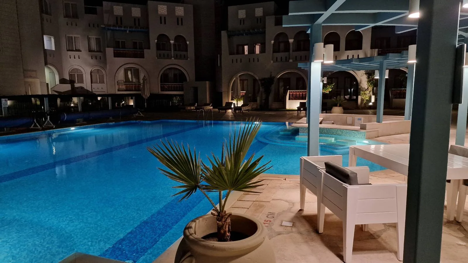 Nighttime view of a large, illuminated outdoor swimming pool surrounded by buildings with arched windows and doors; some poolside furniture and potted plants are visible in the foreground.