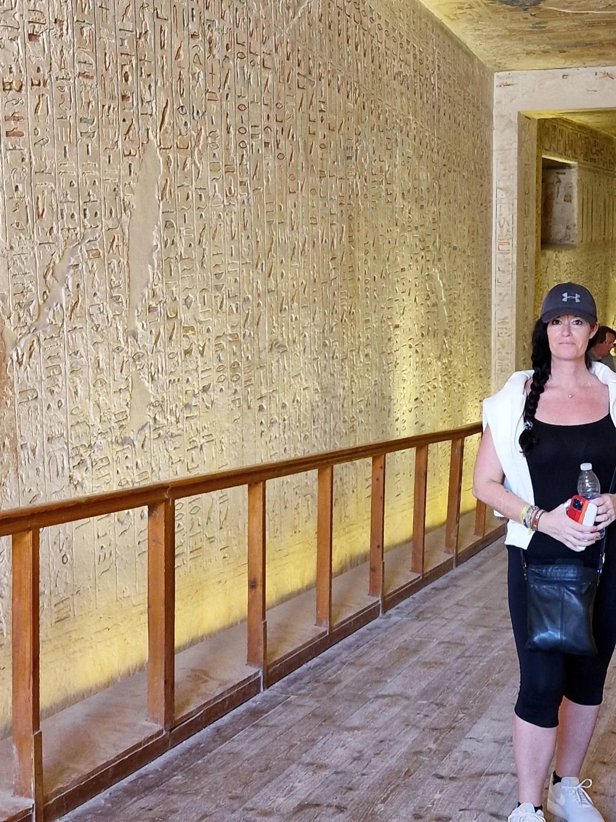 A woman in athletic wear and a cap walks through an ancient corridor with hieroglyphs on the wall, holding a drink bottle.