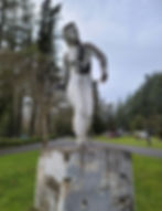 Weathered statue of a figure posing atop a graffitied pedestal in a park setting.