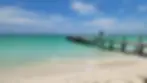 Person walking on a wooden pier over turquoise water beside a sandy beach under a bright blue sky
