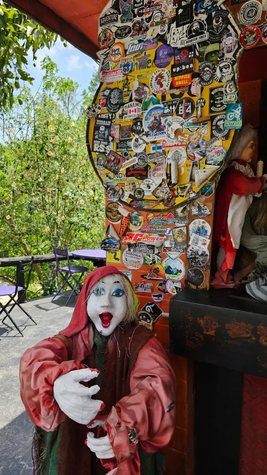 Creepy doll in red hood, outstretched hands in a sticker-covered cafe. Outdoor seating with green foliage in the background.