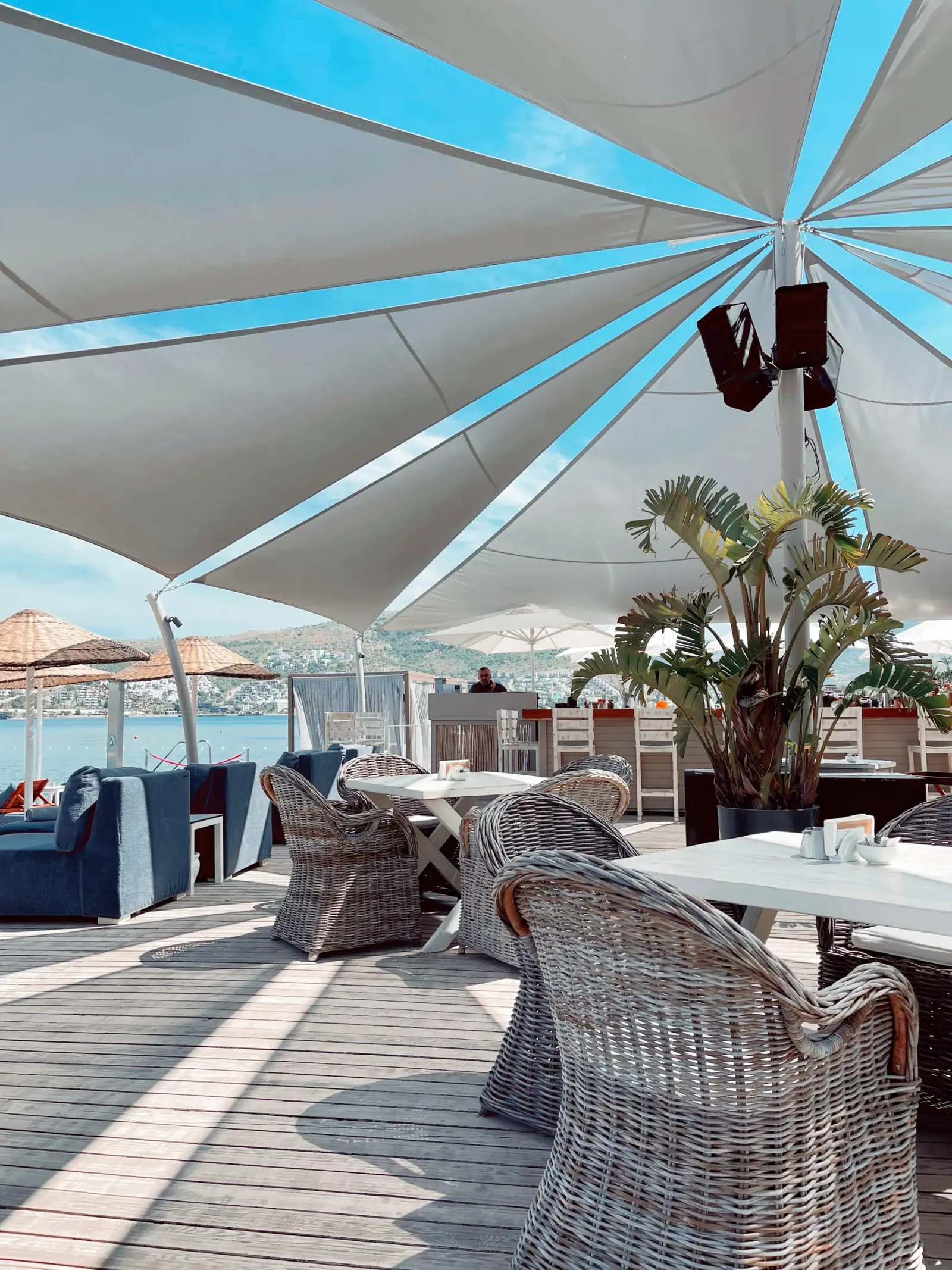 Outdoor seating area with wicker chairs and tables under a large white canopy. The area overlooks water, with additional beach umbrellas and plants nearby.