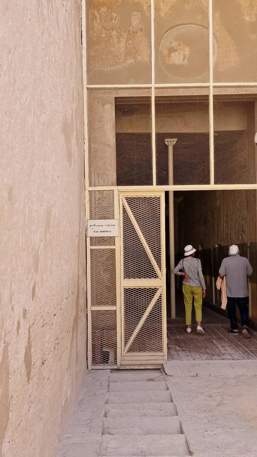 Two people in light hats enter a historical building with high, decorated walls. The stone wall on the left has a weathered appearance. The doorway is framed by a grid-like structure, and the floor is made of large tiles.