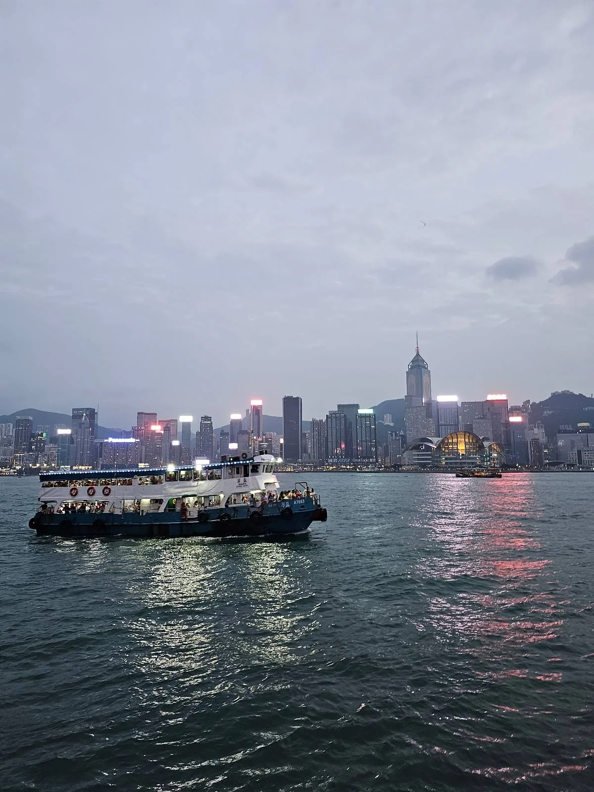 A ferry sails on a body of water with a city skyline and brightly lit skyscrapers in the background during twilight.