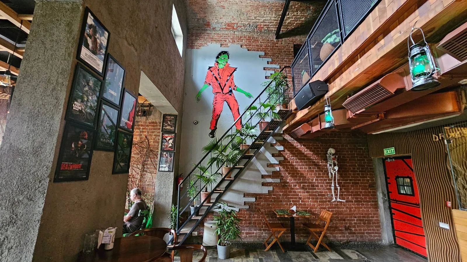 A room with exposed brick walls features a staircase decorated with a green alien figure wearing a red outfit. Framed pictures and posters adorn the walls. Below, two chairs and a small table sit near a skeleton figure near a red door.