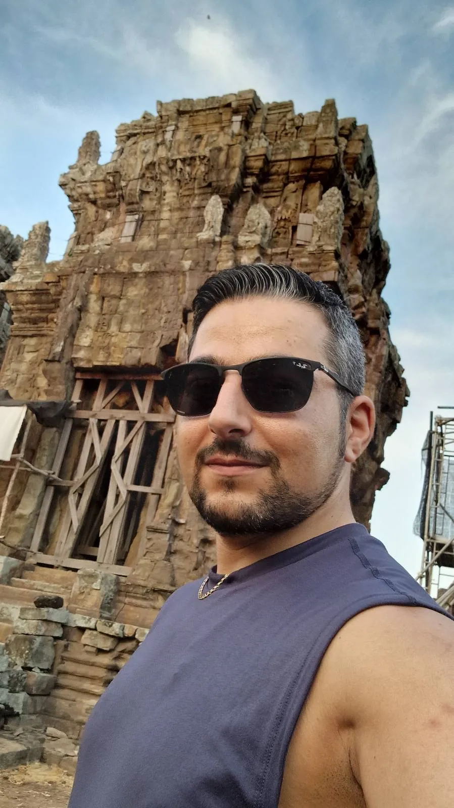 A man wearing sunglasses and a sleeveless shirt takes a selfie in front of an ancient stone temple with intricate carvings, under a partly cloudy sky.