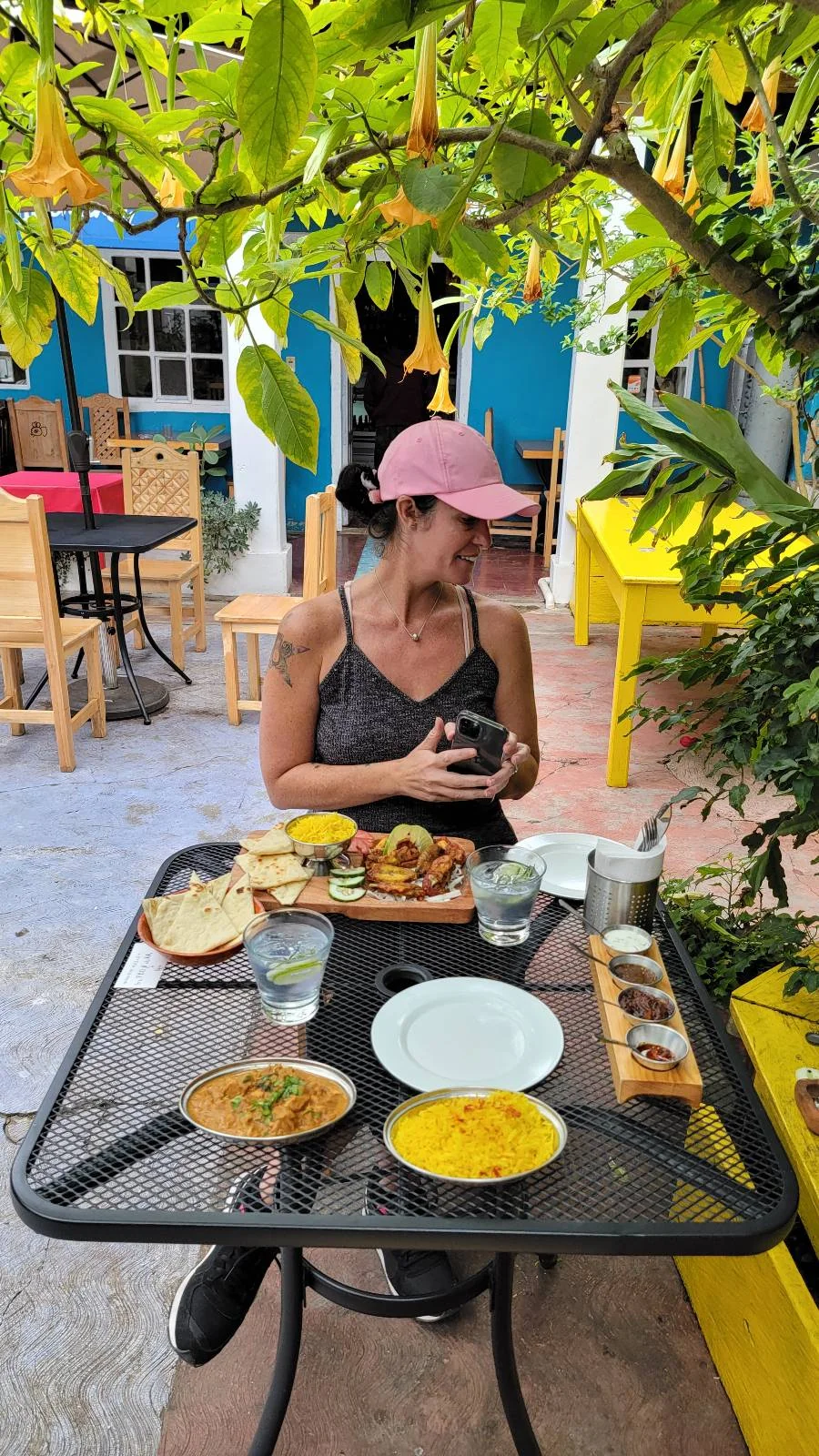 A woman in a pink cap sits at an outdoor cafe table with various dishes, including hummus, olives, salads, and bread. The setting is vibrant, with yellow and blue decor and lush greenery overhead.