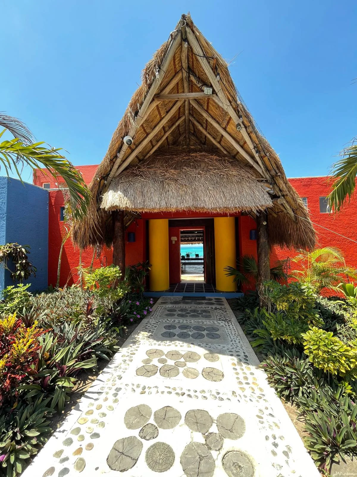 A tropical resort entrance with a thatched roof, vibrant red walls, and colorful designs. A stone pathway leads to an open doorway, revealing a glimpse of the ocean in the background. Lush greenery lines the path under a clear blue sky.
