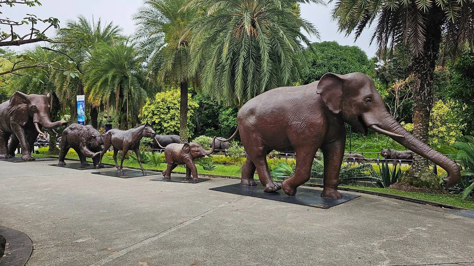 A line of bronze elephant statues, including adults and calves, stands on a path surrounded by lush greenery and palm trees. The scene is outdoors on a cloudy day.
