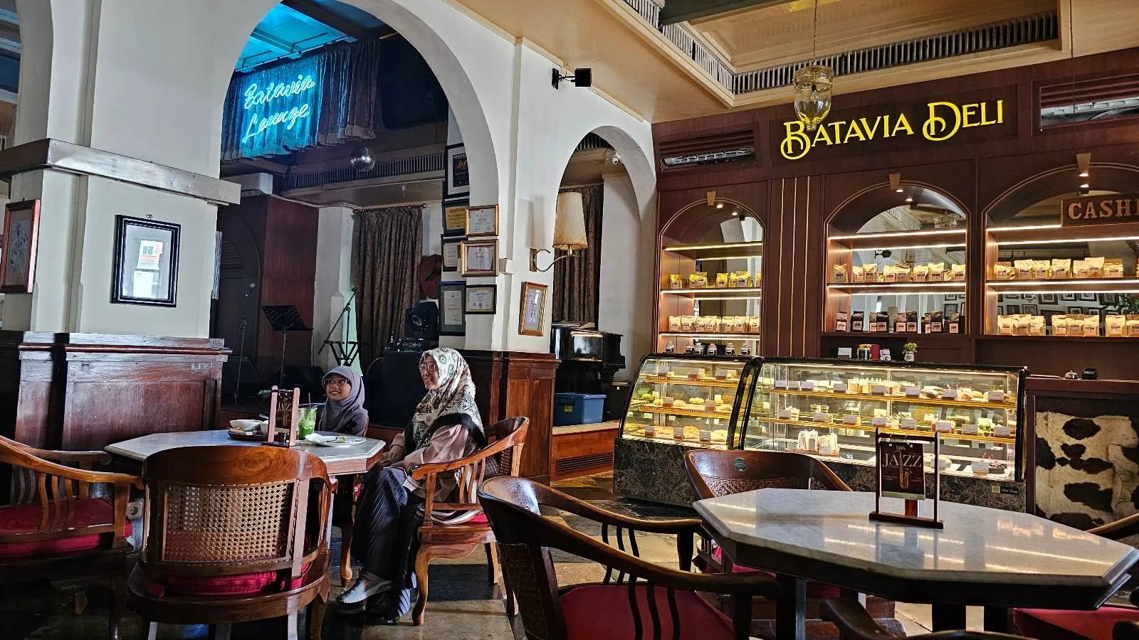 A cozy café with a wooden interior features a "Batavia Deli" sign above a display filled with pastries. Two people sit at a table, and nearby, a neon sign glows on the wall. The setting is warmly lit and inviting.
