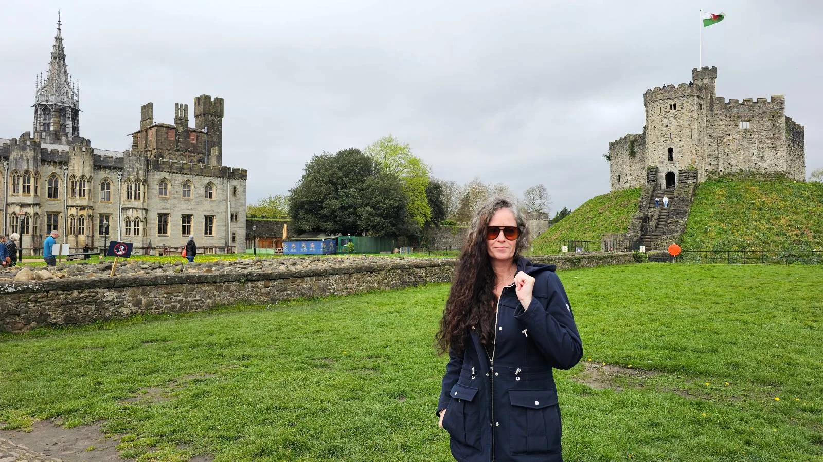 Woman wearing sunglasses and a dark coat standing in front of a historic castle with green lawns.