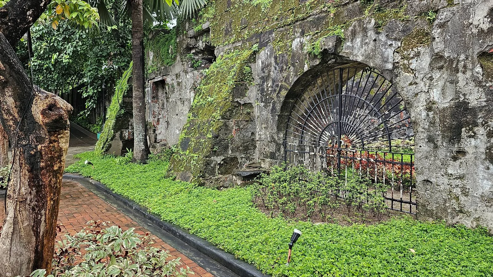 A moss-covered stone wall with a large arched iron gate, surrounded by green plants and trees, lines a brick pathway in a lush garden setting.