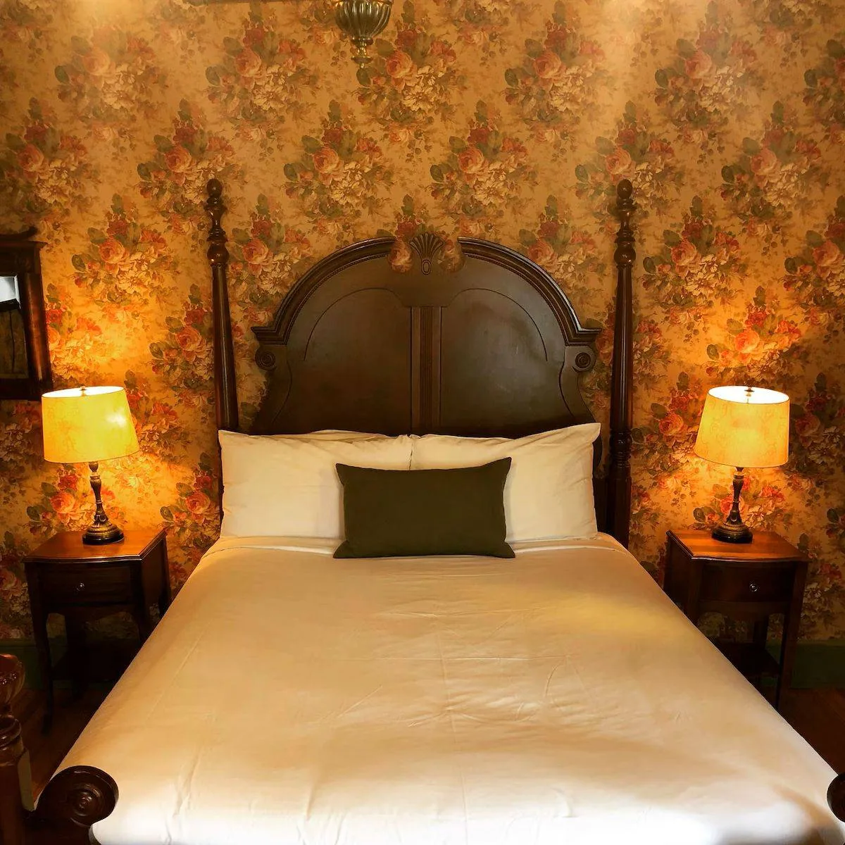 A cozy bedroom features a bed with a dark wooden headboard and white bedding. There's a green accent pillow in the center. Two bedside tables, each with a lamp, flank the bed. The wallpaper has a floral pattern with warm colors.