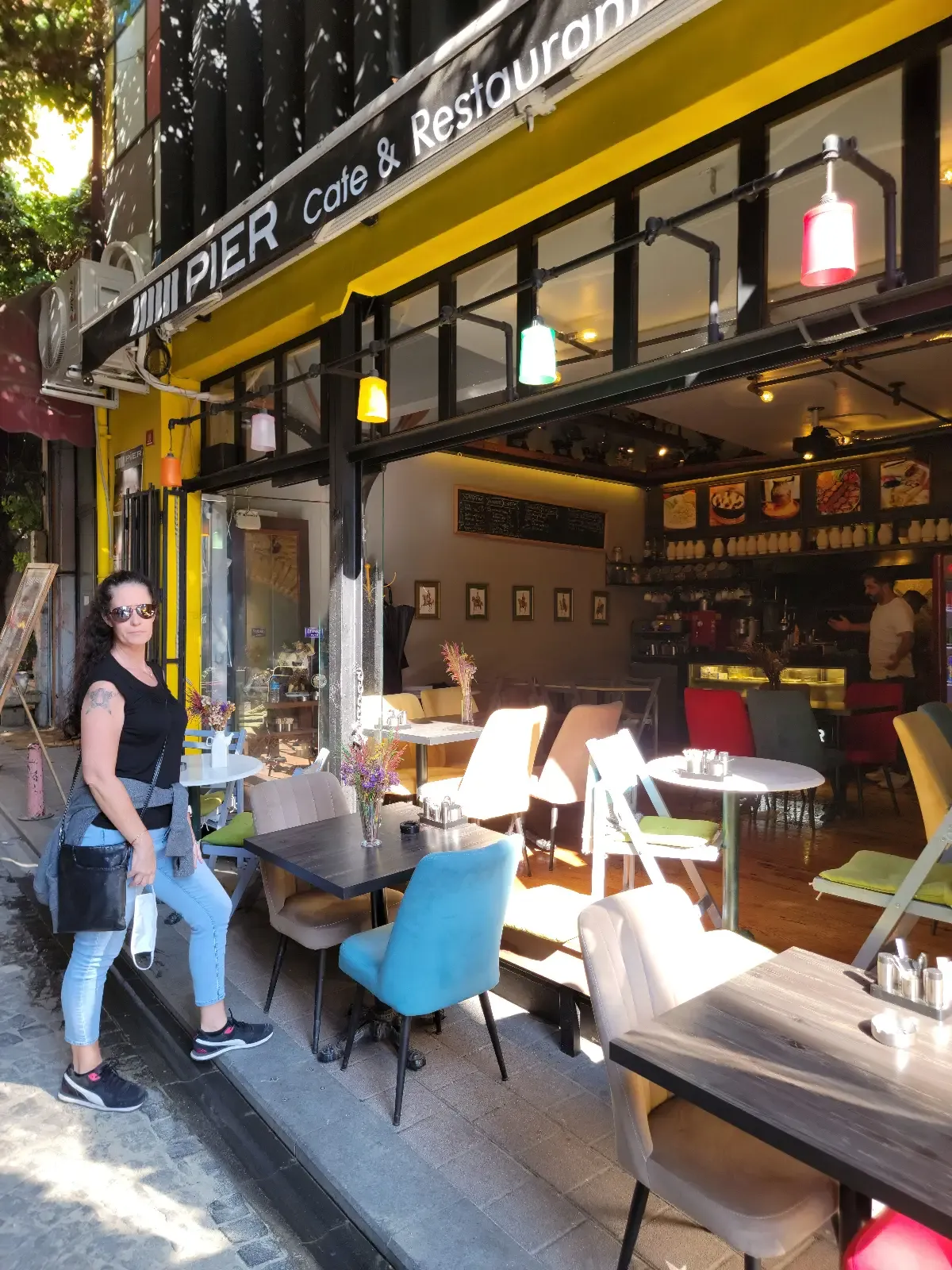 A woman stands outside a colorful café with modern chairs and tables set up on the sidewalk. The café has large windows, hanging lights, and a sign reading "Coffee Bar." Sunlight casts shadows on the street.