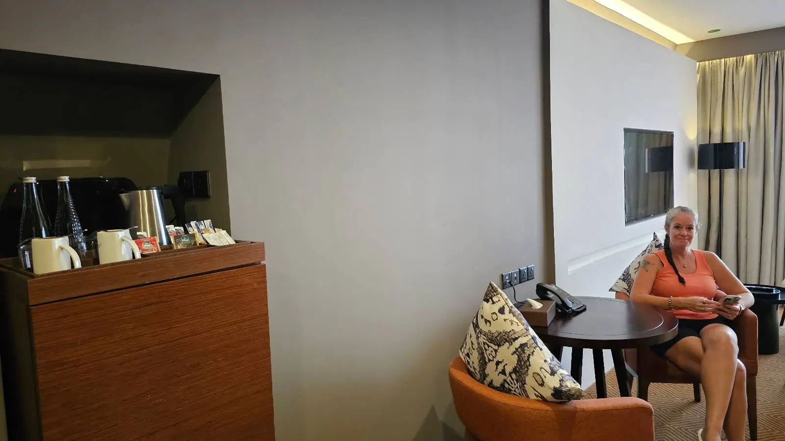 A person is sitting on an orange chair at a round table in a modern, well-lit room. There's a wall-mounted TV and a small cabinet with a kettle and cups on the left. A large window with curtains is in the background, adding a cozy ambiance.