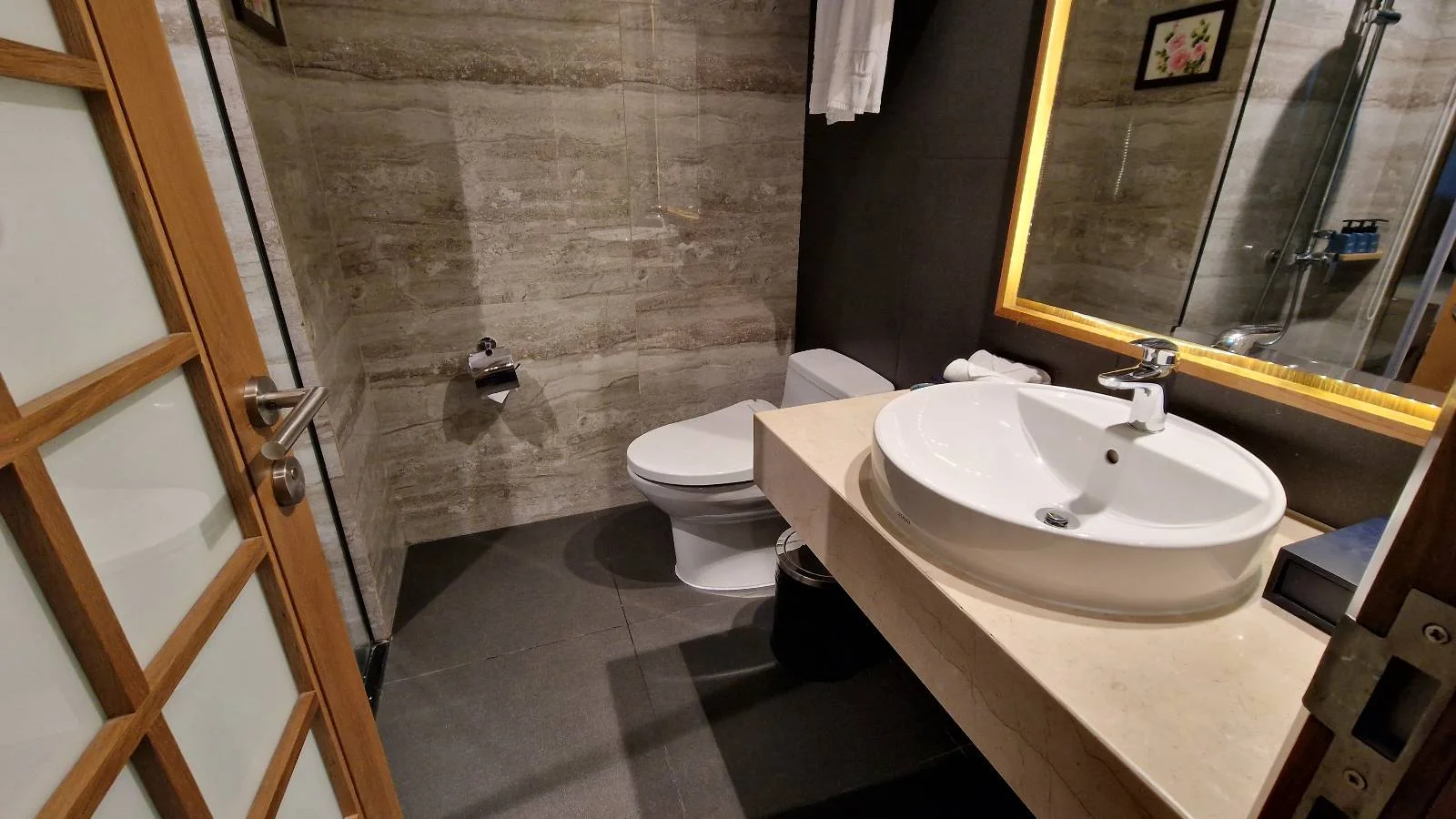 A modern bathroom with a toilet and a round sink on a beige countertop. The walls are tiled in a light gray stone texture, and a large mirror with a lighted frame hangs above the sink. The floor is dark gray.