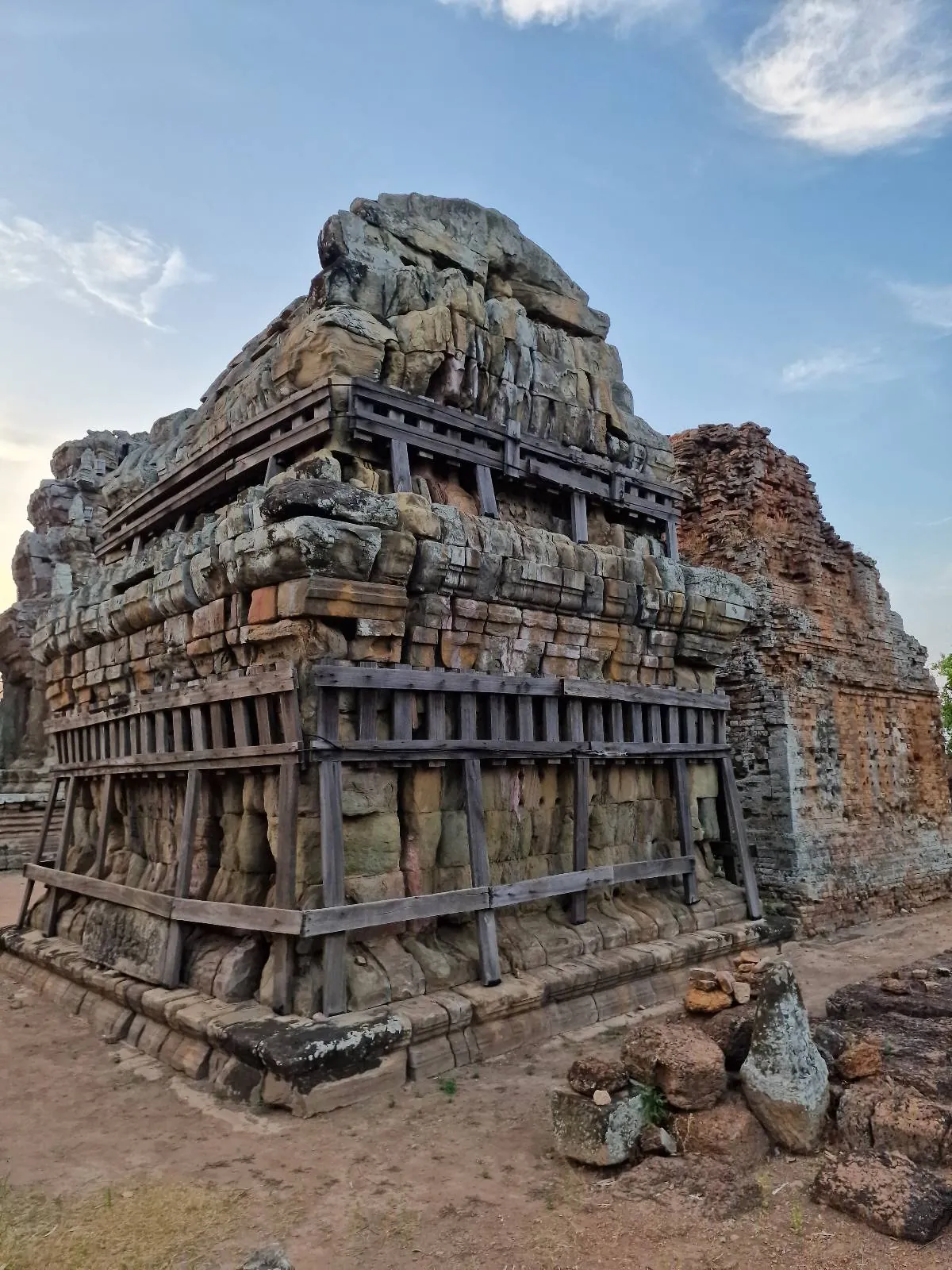 An ancient stone structure with intricate carvings and wooden supports stands under a blue sky with scattered clouds, surrounded by rocky terrain and sparse vegetation.