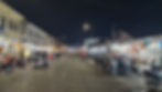 Night market scene with street vendors, motorcycles, and tuk-tuks. Bright lights illuminate shops and people. Calm, bustling atmosphere.