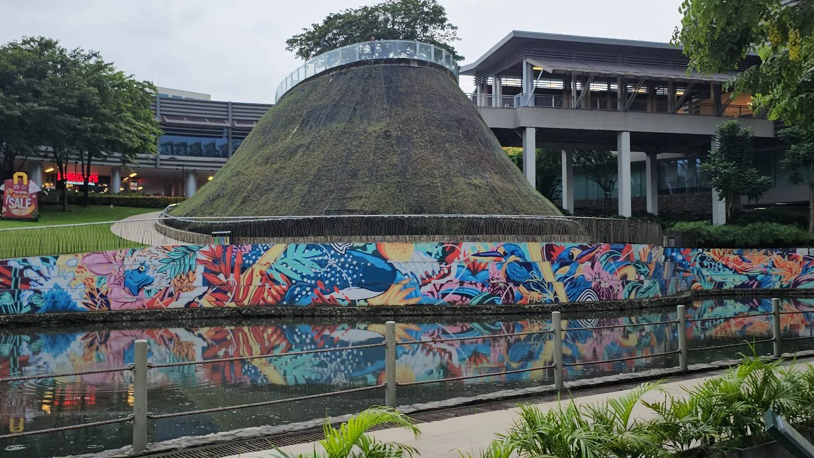 A vibrant mural with abstract designs adorns a wall along a canal. Nearby, a grass-covered, conical structure stands next to a modern building. Trees and foliage surround the area, adding a natural touch to the urban scenery.