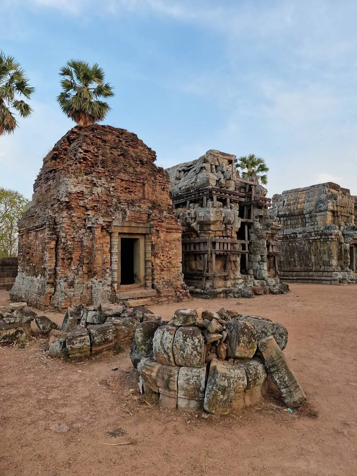 An ancient stone temple with a weathered entrance and palm trees in the background, surrounded by ruins and sandy ground under a blue sky.